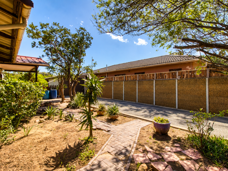 A rear exterior view featuring a paved walkway, established trees, and a precast concrete boundary wall alongside the main house structure.