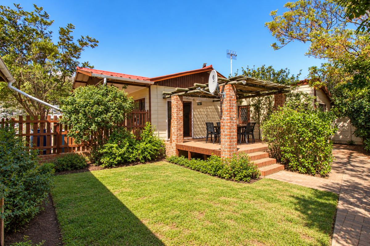 A well-maintained rear garden featuring a brick patio with a timber pergola, surrounded by established greenery and a paved walkway.