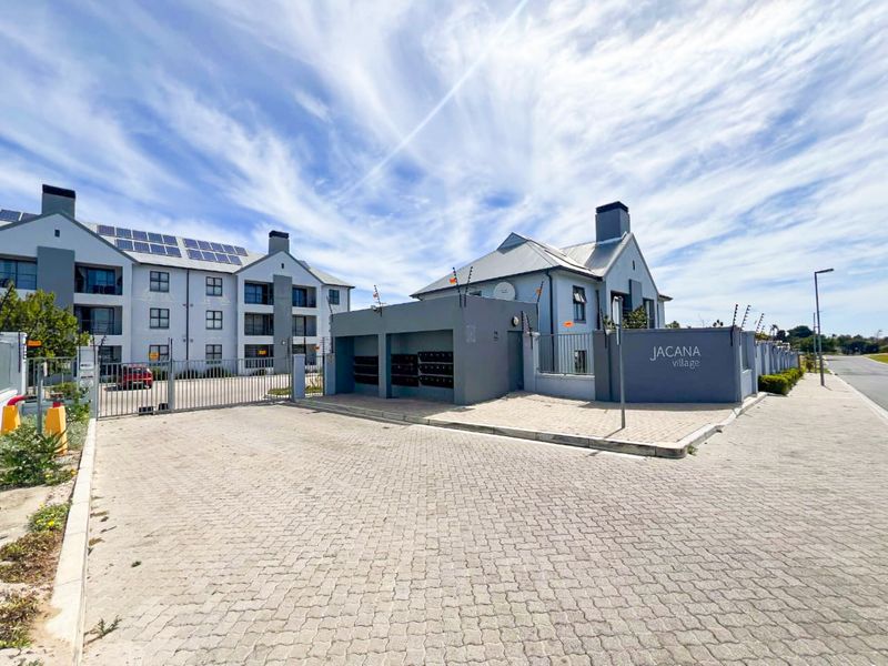 Front entrance view of Jacana Village apartment complex featuring paved driveway, security gate, post boxes, and solar panel installations on the roof.