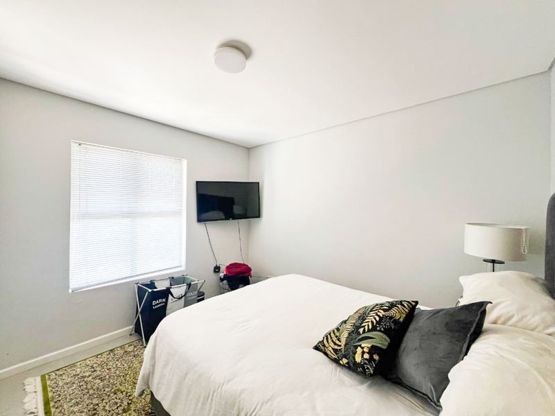 A well-lit bedroom featuring neutral walls, a window with fitted blinds, and a wall-mounted television.