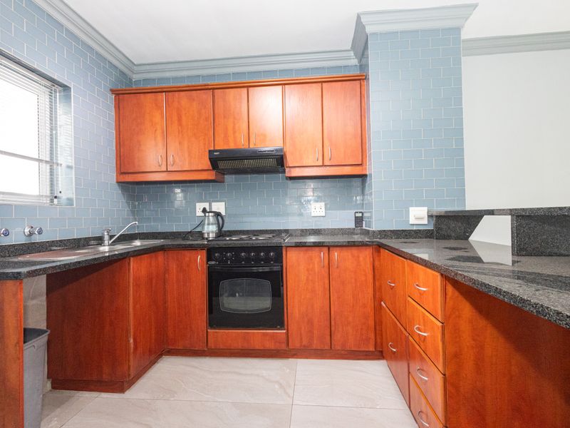 A functional kitchen featuring granite countertops, wooden cabinetry, built-in oven, hob, extractor fan, and blue tiled backsplash with neutral floor tiles.