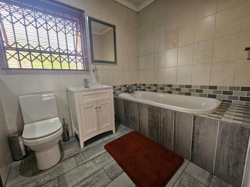 A well-maintained bathroom featuring a built-in vanity, bathtub, toilet, and modern tiled finishes with a security gate on the window.