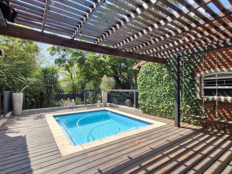 A well-maintained outdoor pool area featuring a wooden deck, a pergola structure, and a brick wall, creating a pleasant space for relaxation.