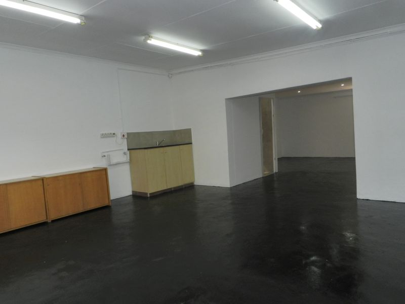 An empty open-plan space featuring painted concrete floors, fluorescent lighting, and a basic kitchenette area with built-in cabinetry and a sink.