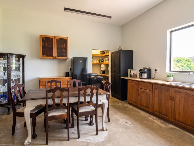 A functional kitchen featuring wooden cabinetry, a stainless steel sink, and an adjoining pantry, with a dining area and concrete flooring.