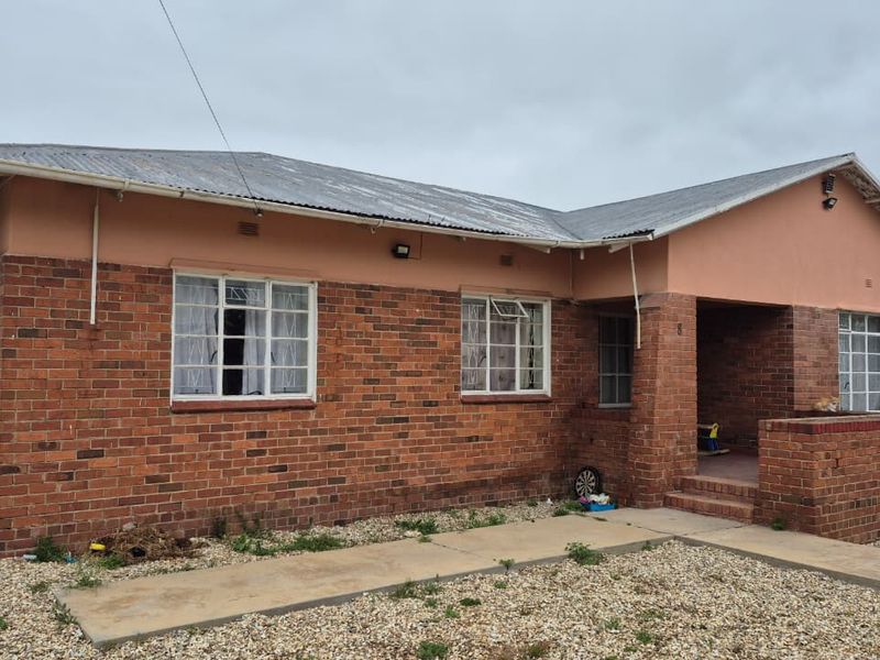Front elevation of a single-story brick house featuring a covered entrance porch, security-barred windows, and a corrugated metal roof.