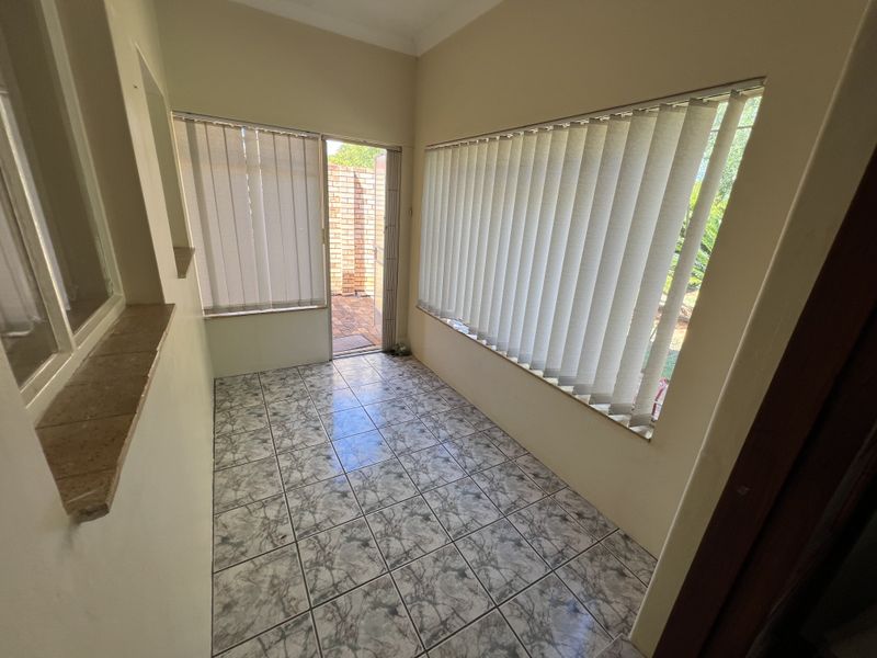 A sunlit, empty enclosed patio area featuring tiled flooring, large windows with vertical blinds, and direct access to the outdoor space.