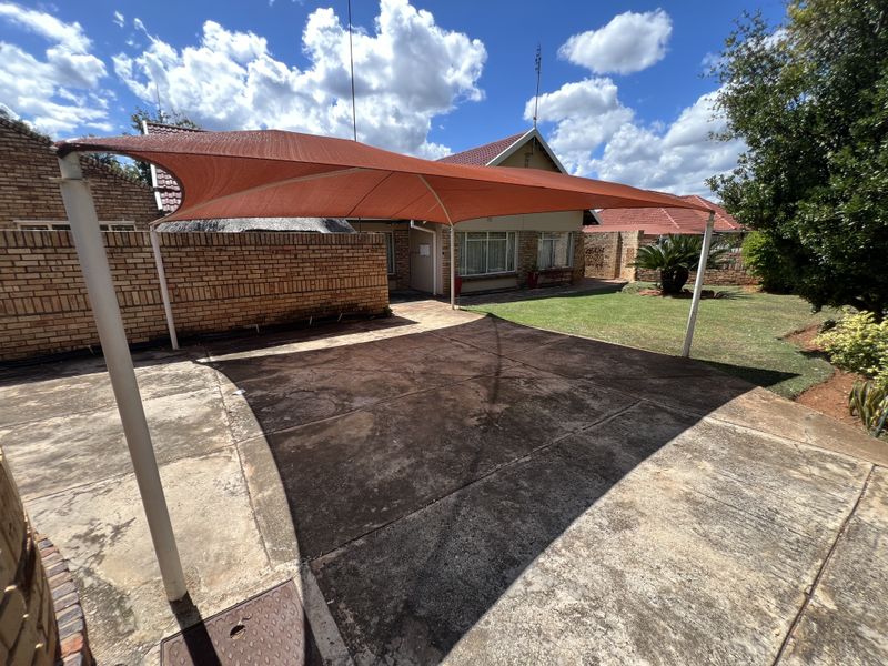 Rear exterior view featuring a large shade sail over a concrete paved area, brick boundary walls, and a well-maintained lawn.