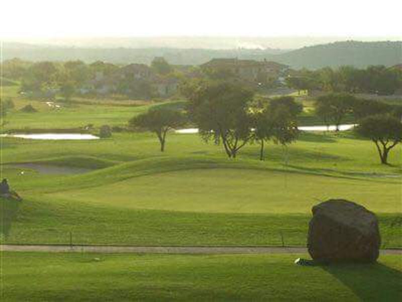 A scenic view overlooking a well-maintained golf course with rolling green fairways, mature trees, and a water feature in the distance.