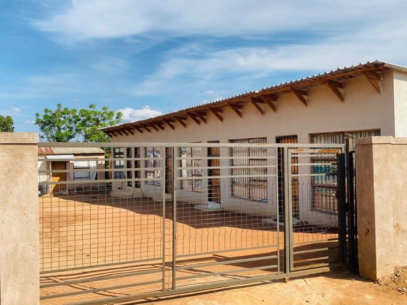 Front view of a single-story house featuring a metal security gate, perimeter wall, and steel-framed windows under a corrugated metal roof.