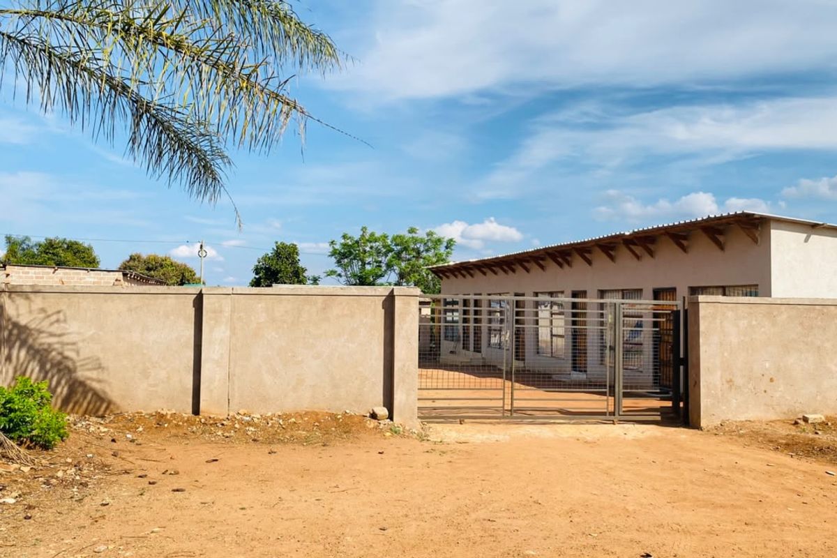 Front view of a single-story property featuring a perimeter wall, metal gate, and a corrugated metal roof with security bars on the windows.
