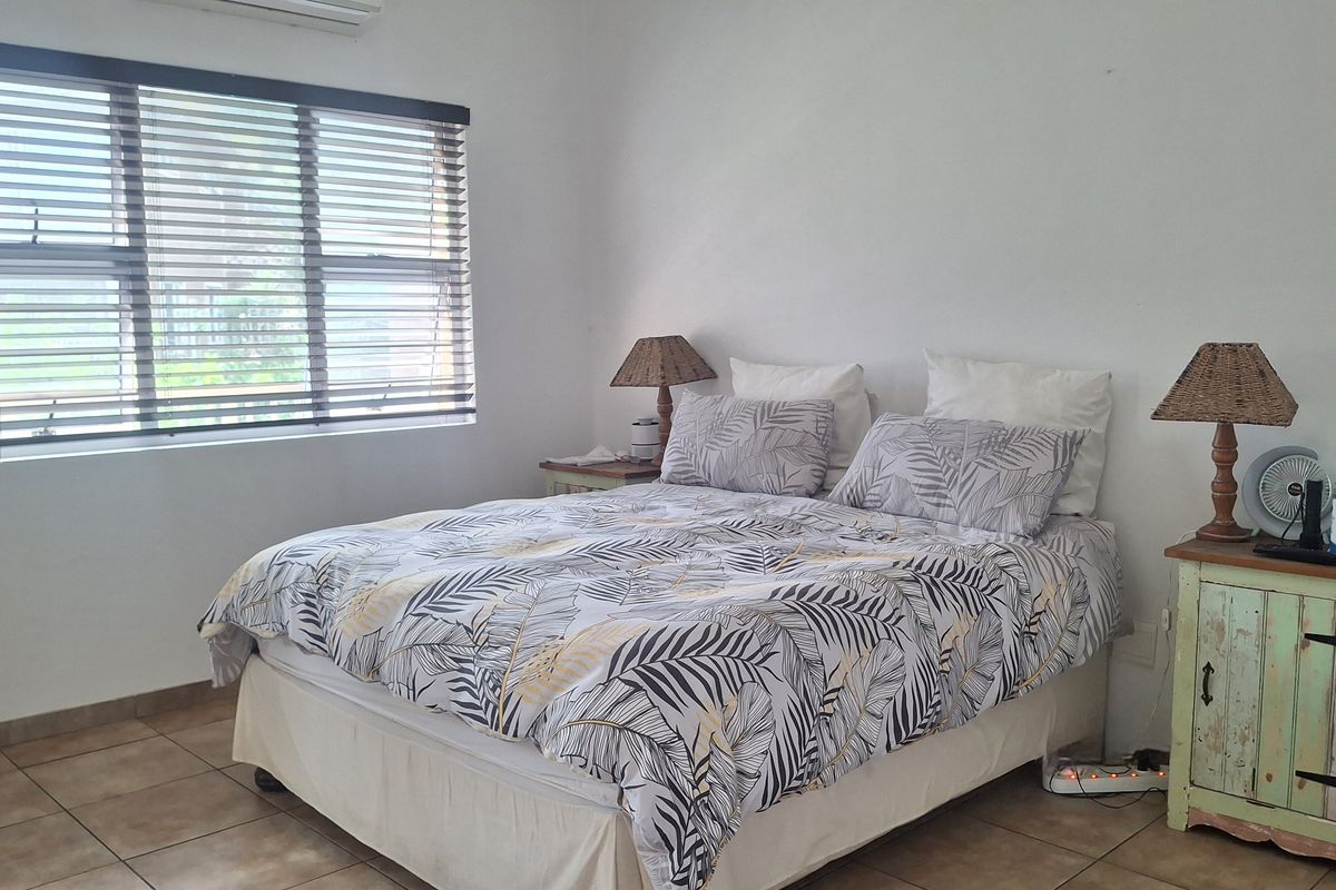 A functional bedroom featuring tiled flooring, a wall-mounted air conditioning unit, and large windows fitted with venetian blinds.