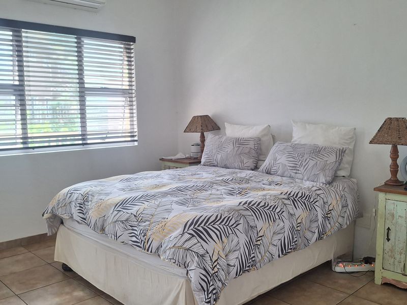 A functional bedroom featuring tiled flooring, a wall-mounted air conditioning unit, and large windows fitted with venetian blinds.