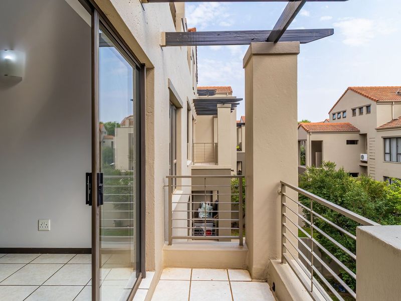 A clean, tiled balcony featuring a timber pergola, metal balustrade, and sliding glass door access, overlooking the apartment complex grounds.