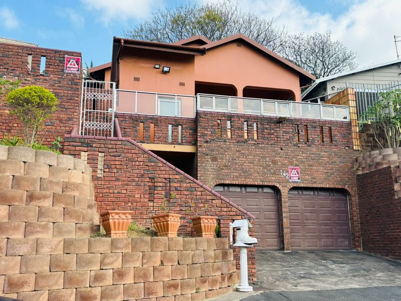 Front elevation of a multi-level face brick house featuring a double garage, elevated balcony with glass balustrade, and tiered retaining wall.