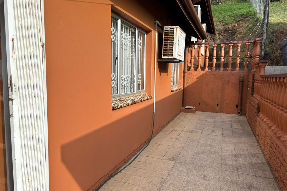 A paved rear patio area featuring a balustrade, security gates on windows, and an exterior-mounted air conditioning unit.