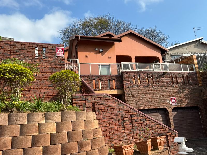 Front elevation of a multi-level face brick house featuring a double garage, elevated balcony with glass balustrades, and a tiered retaining wall.