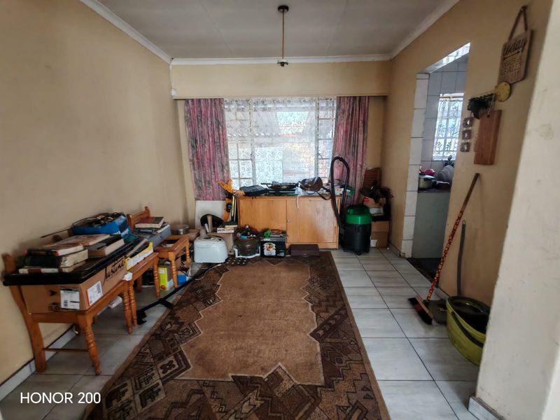 A living room area featuring tiled flooring and window security bars, leading into an adjacent kitchen space. The room requires cosmetic updates and maintenance.