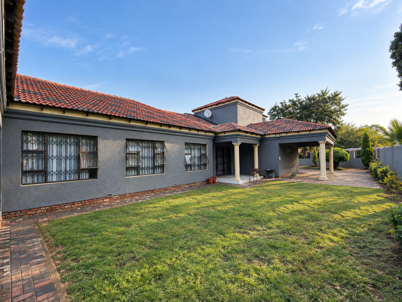 Rear view of a well-maintained house featuring a covered patio, terracotta tiled roof, and a spacious lawn with brick paved pathways.
