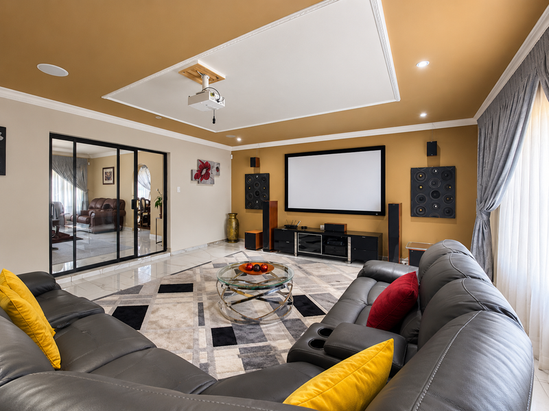 A dedicated cinema room featuring a ceiling-mounted projector, large fixed projection screen, and sleek sliding glass doors leading to an adjacent living space.