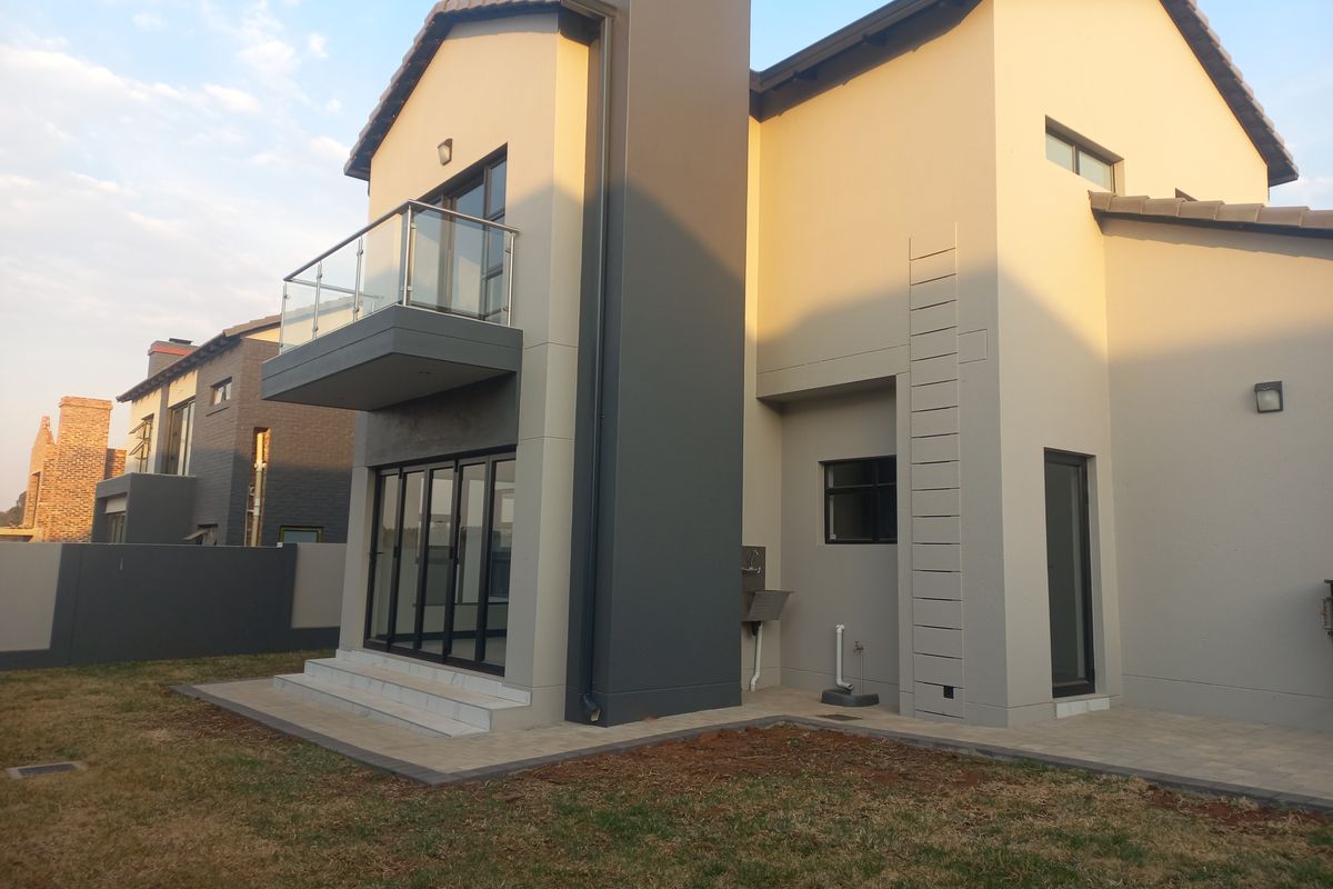 Rear view of a modern double-story house featuring a balcony with glass balustrade, sliding doors, and a paved patio area.