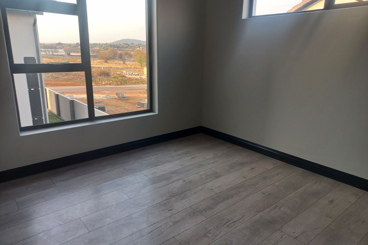 A bright, empty bedroom featuring modern laminate flooring, dark skirting boards, and large aluminium-framed windows with views of the surrounding landscape.