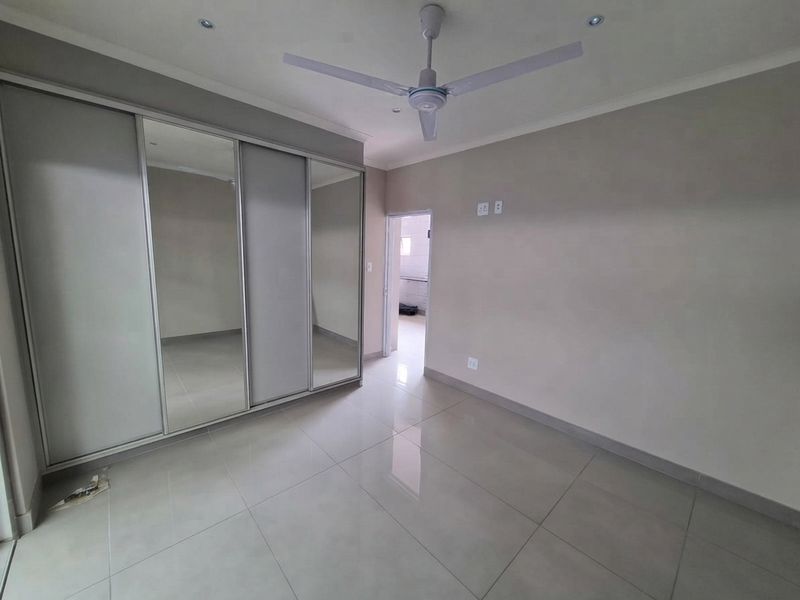 A clean, empty bedroom featuring large mirrored built-in cupboards, neutral tiled flooring, a ceiling fan, and recessed lighting.