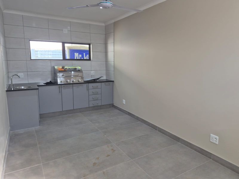 A clean, empty scullery or entertainment prep area featuring built-in braai, granite countertops, stainless steel sink, and modern grey cabinetry.