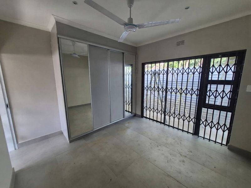 A bright, empty bedroom featuring tiled flooring, a large built-in mirrored wardrobe, a ceiling fan, and a secure sliding door with a metal security gate.