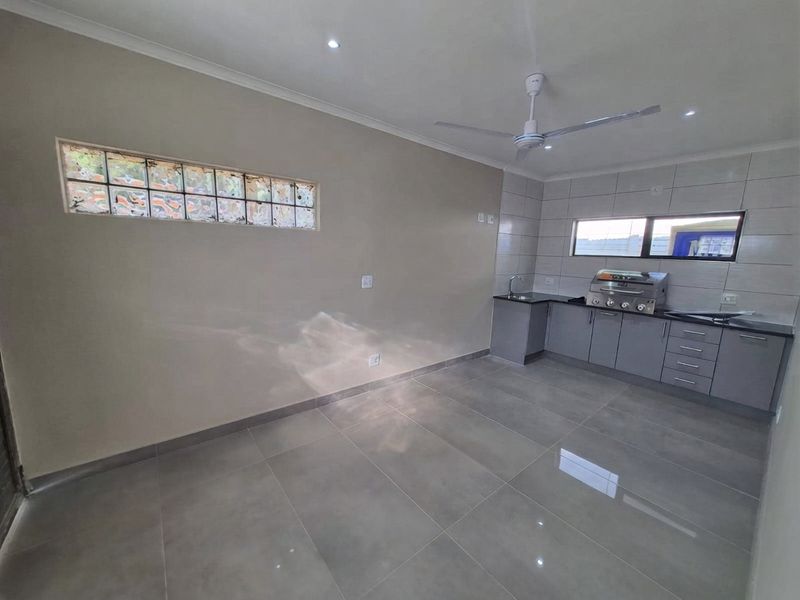 A modern, empty kitchen featuring grey cabinetry, stone countertops, a built-in gas braai, tiled flooring, and a unique glass block window.