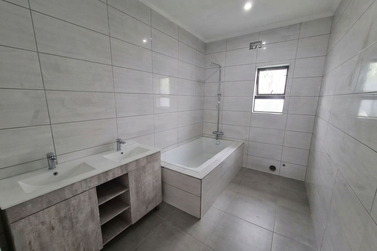 Modern bathroom featuring a double vanity with wood-look cabinetry, a built-in bathtub with shower mixer, and neutral floor-to-ceiling wall tiles.