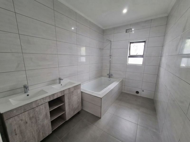 Modern bathroom featuring a double vanity with wood-look cabinetry, a built-in bathtub with shower mixer, and neutral floor-to-ceiling wall tiles.