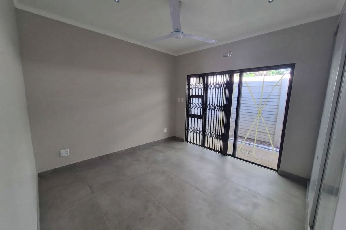 A clean, empty bedroom featuring neutral tiled flooring, a ceiling fan, and a sliding glass door with a security gate leading to the exterior.
