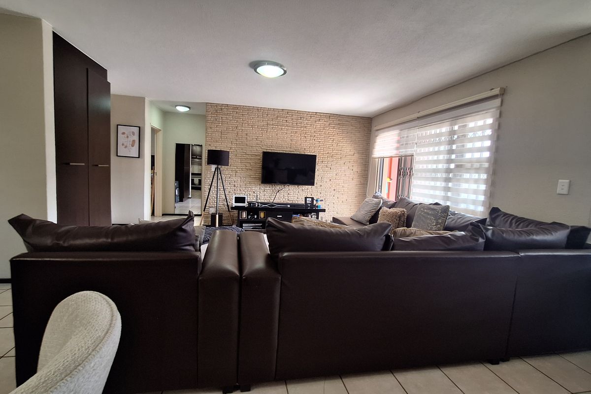 Spacious living room featuring an exposed brick accent wall, tiled flooring, and large windows with roller blinds, offering a comfortable and functional layout.