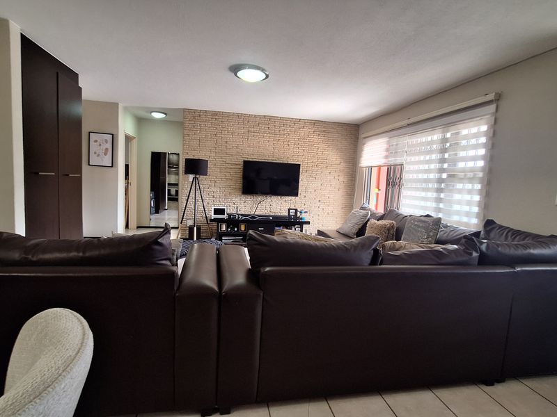 Spacious living room featuring an exposed brick accent wall, tiled flooring, and large windows with roller blinds, offering a comfortable and functional layout.