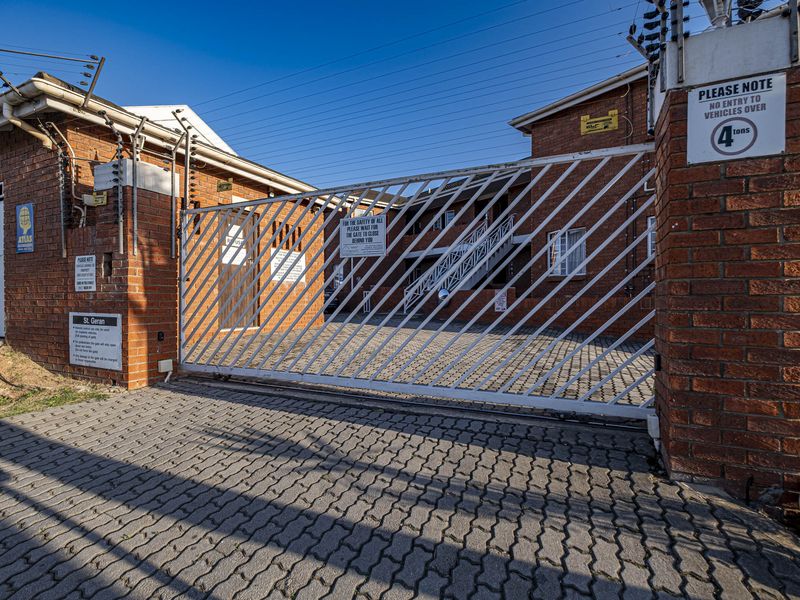Front entrance view of the apartment complex featuring a secure sliding gate, brick perimeter wall, and electric fencing for added security.