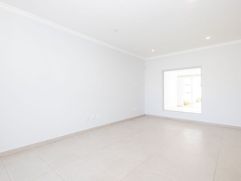 A bright, empty living room featuring neutral tiled flooring, recessed downlighting, and clean white walls, offering a versatile space ready for furnishing.