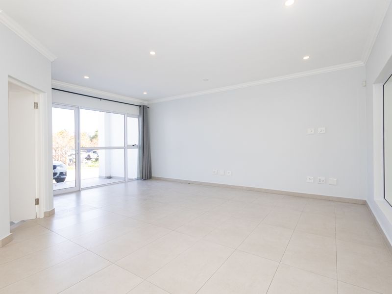 A bright, spacious, and empty living room featuring neutral tiled flooring, recessed downlights, and a large sliding glass door providing natural light.