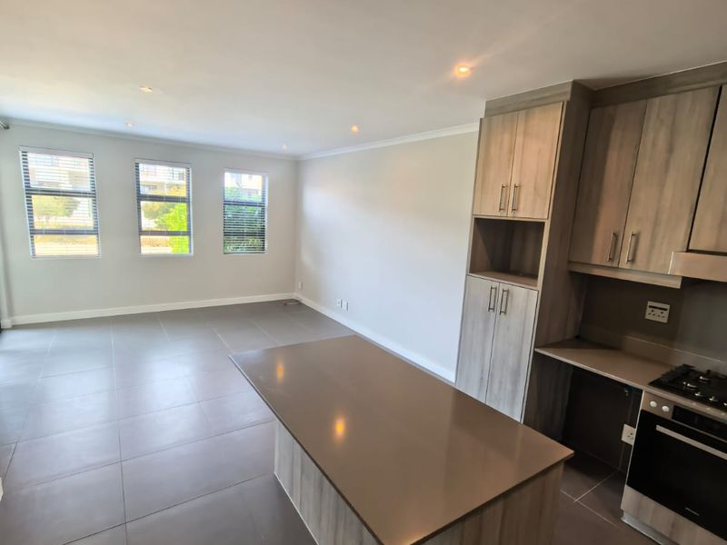 Spacious open-plan living and kitchen area featuring modern wood-finish cabinetry, a central island with stone countertop, built-in oven, and large tiled flooring.
