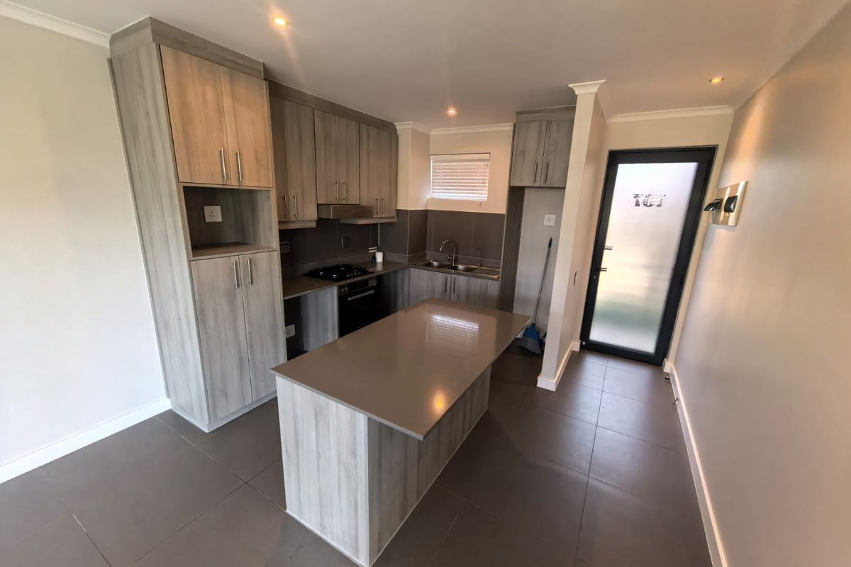 Modern kitchen featuring light wood-grain cabinetry, a central island with stone countertop, built-in oven and hob, and tiled flooring throughout.