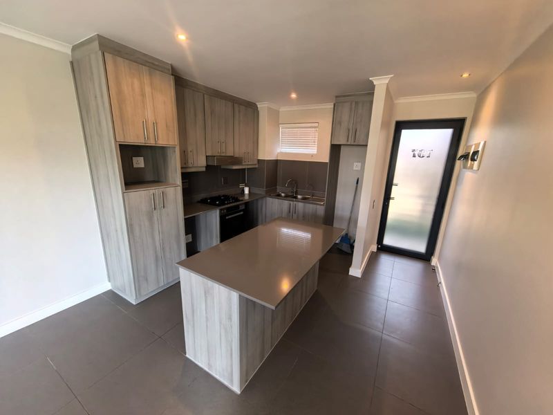 Modern kitchen featuring light wood-grain cabinetry, a central island with stone countertop, built-in oven and hob, and tiled flooring throughout.