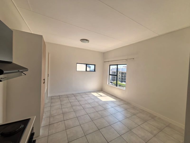 A bright, empty open-plan living and kitchen area featuring neutral tiled flooring, built-in cabinetry, and large windows providing natural light.