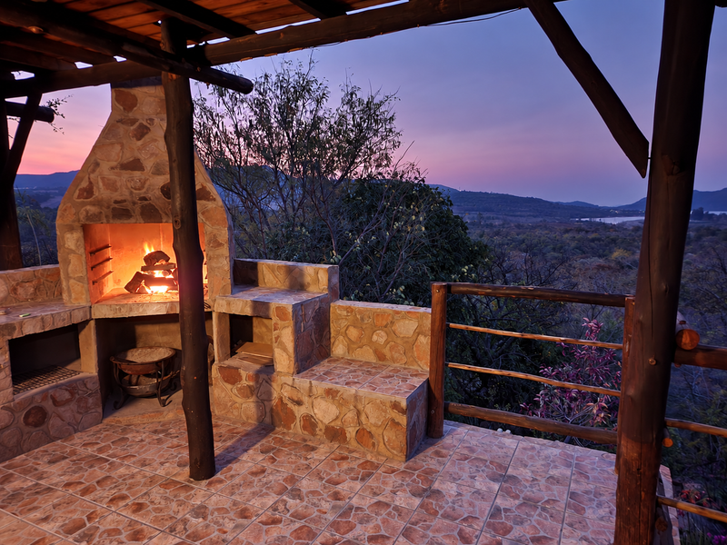 Covered outdoor patio featuring a stone-clad built-in braai, tiled flooring, and timber structure, offering scenic elevated views of the surrounding landscape.