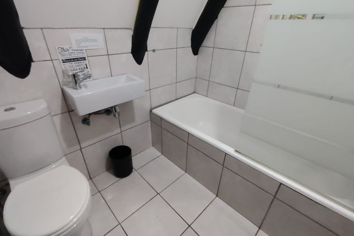A compact bathroom featuring a built-in bathtub with a glass screen, wall-mounted basin, and toilet, finished with neutral floor and wall tiling.