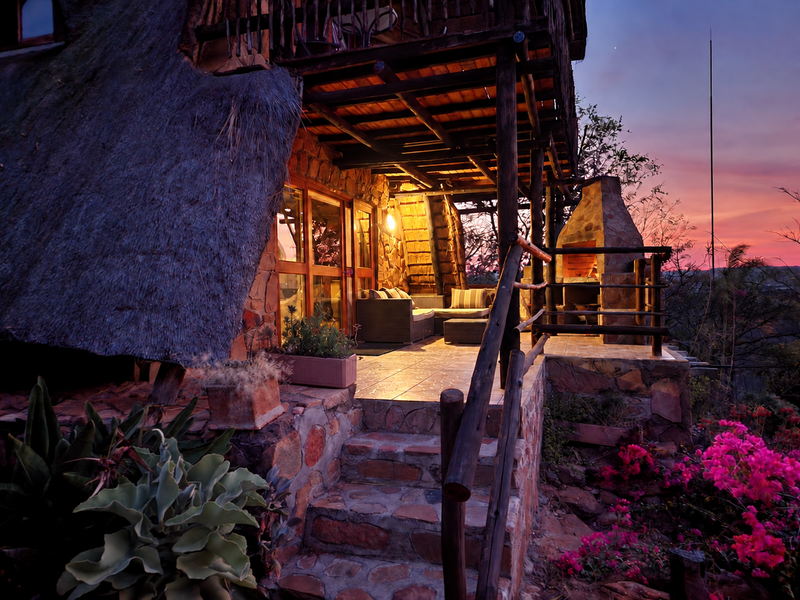 A charming rustic patio featuring stone cladding, a built-in fireplace, and a thatched roof, offering a cozy outdoor living space with sunset views.