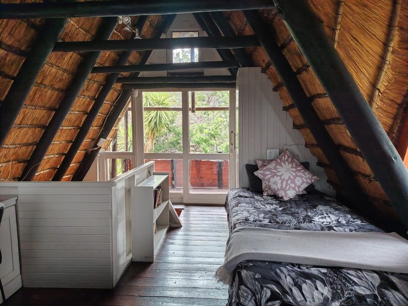 A cozy loft-style bedroom featuring exposed timber rafters, a traditional thatched roof, timber flooring, and direct access to an outdoor balcony.
