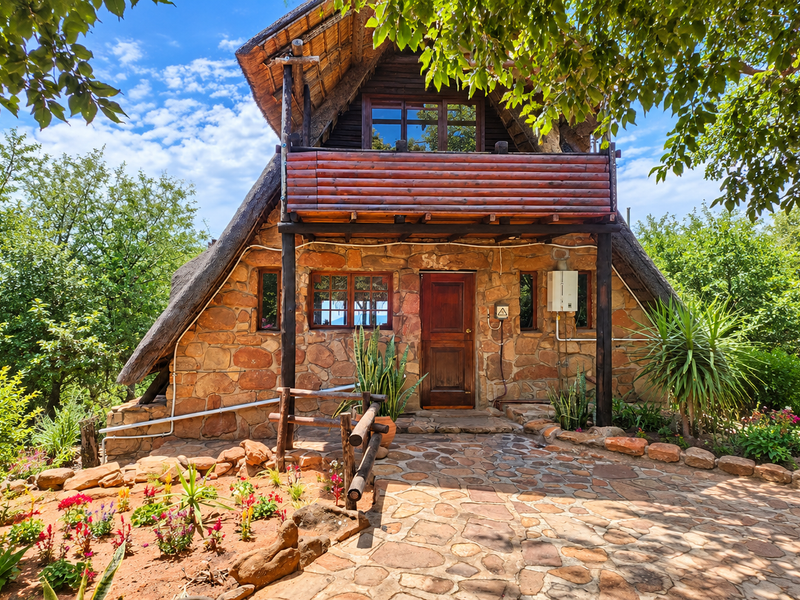 A charming A-frame cottage featuring rustic stone cladding, a thatched roof, and a timber balcony, set within a lush, well-maintained garden.