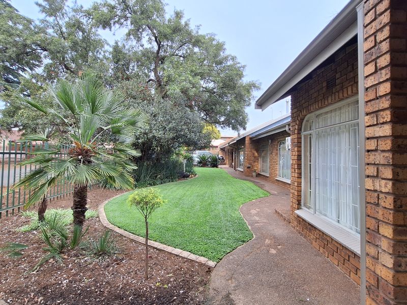 A well-maintained front garden featuring a manicured lawn, paved walkway, and face brick exterior with secure window bars and metal perimeter fencing.