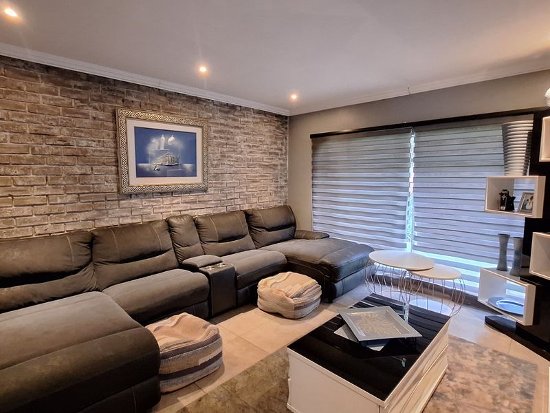 A comfortable living room featuring a textured exposed brick feature wall, modern recessed downlighting, and large windows fitted with adjustable roller blinds.