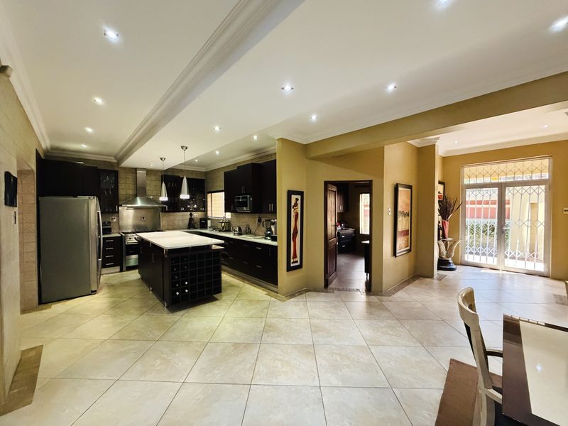 Spacious open-plan kitchen featuring a central island, dark cabinetry, stainless steel appliances, and tiled flooring, leading to a bright dining area with garden access.
